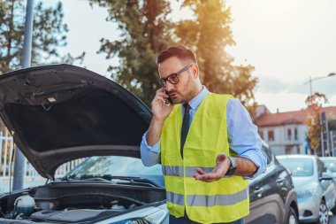 Yansıtıcı güvenlik yeleği giyen bir adam açık araba kaputunun yanında duruyor ve bir araç arızası nedeniyle yol kenarında yardım çağrısı yapıyor..