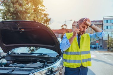 Emniyet yeleği giyen bir adam açık araba kaputunun yanında duruyor, hayal kırıklığına uğramış görünüyor, şehir merkezinde yol yardımı için telefon ediyor..