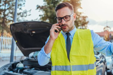 Güvenlik yeleği giyen bir adam, güneşli bir günde araba arızasından sonra yol yardımı istiyor. Açık araba kaputu motor arızası gösteriyor..