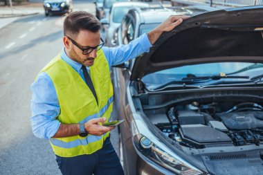 Yansıtıcı yelek giyen bir adam, güneşli bir günde yol kenarındaki cep telefonundan yardım isterken bir araba motoru arızasını inceliyor..