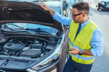 Yansıtıcı yelek giyen profesyonel tamirci kaputu açık araba motorunu inceliyor. Yol yardımı ve araç bakımı kavramı. Otomobili incelerken telefonu tutan adam.