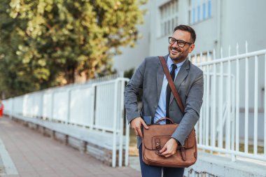 İş adamı gülümsüyor ve şehir kaldırımında deri bir çantayla yürüyor. Profesyonel ve kendinden emin, takım elbiseli ve kravatlı, kentsel bir iş ortamı yaratıyor..