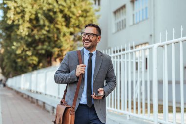 Takım elbiseli kendine güvenen bir işadamı elinde telefon ve çantayla açık havada yürür. Parlak bir günde profesyonellik ve başarı sergilerler..