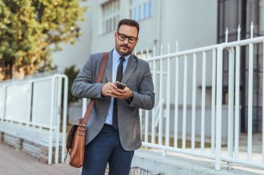 Ofis binasının yanında yürürken telefonunu kontrol eden gözlüklü profesyonel bir işadamı. Deri bir çanta taşıyor ve takım elbise giyiyor. Kurumsal bir yaşam tarzını temsil ediyor..