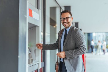 Gülümseyen bir işadamı, ATM 'yi kullanarak bir şehir bölgesinde para çekiyor. Kendinden emin ve profesyonel görünüyor, güneşli bir günde takım elbise ve kravat giyiyor..