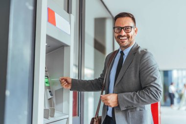 Takım elbiseli ve gözlüklü neşeli bir işadamı ATM 'yi para transferi için kullanıyor. Pozitif tavrı bankacılık hizmetlerindeki güven ve memnuniyeti gösteriyor..
