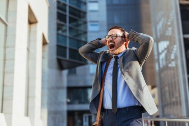 Takım elbiseli stresli işadamı, ofis binasının önünde hayal kırıklığı içinde bağırıyor. Şirket ortamında stres ve endişelerini dile getiriyor..