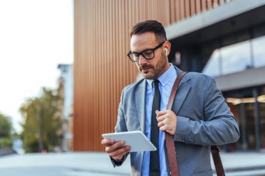 Çağdaş bir ofis binasının önünde dikilirken tablet kullanan takım elbiseli profesyonel bir işadamı iş dünyasındaki teknoloji ve iletişimi aktarıyor..