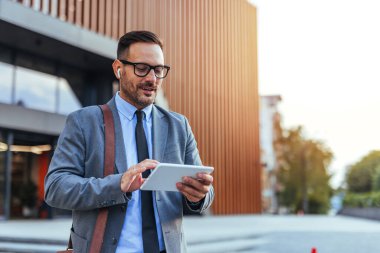 Çağdaş bir ofis binasının önünde dijital tablet kullanan takım elbiseli kendine güvenen bir işadamı. Gözlük ve kulaklık takıyor, modern teknoloji ve profesyonel iletişim sergiliyor..