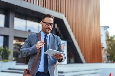 Modern bir ofis binasının önünde yürürken tablet kullanan kendine güvenen işadamı, modern teknoloji ve profesyonel iletişimi örnek alıyor. Takım elbise giyiyor ve deri bir çanta taşıyor..