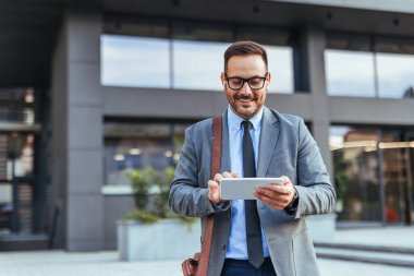 Modern bir şehir ofisinin önünde gülümseyen dijital tablet kullanan takım elbiseli profesyonel bir erkek. İş teknolojisini ve iletişimi temsil eden kendinden emin ve ilgili görünüyor..