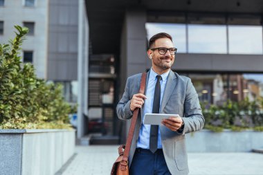 Takım elbiseli ve kravatlı gülümseyen işadamı elinde tabletle modern bir ofis binasının önünde yürüyor. Kendinden emin ve profesyonel görünüyor. Başarılı bir iş ortamı olduğunu gösteriyor..