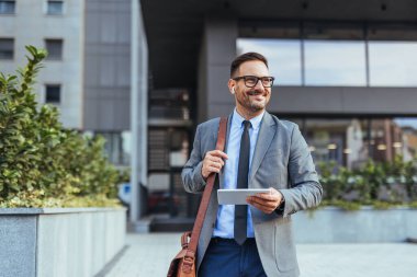Ofis binasının önünde elinde tablet tutarken gülümseyen takım elbiseli kendine güvenen bir işadamı. Başarı, teknoloji ve profesyonel yaşam tarzını temsil eder..
