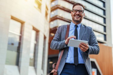 Takım elbiseli gülümseyen işadamı, ofis binasının önünde tablet kullanıyor, modern çalışma ortamında güven ve başarı yayıyor..