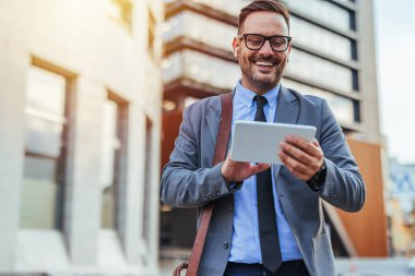 Takım elbiseli ve gözlüklü kendinden emin bir erkek dışarıda dururken teknolojinin tadını çıkarıyor. Modern kentsel ortam iş hayatı ve bağlantıyı gözler önüne seriyor.
