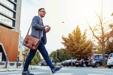 Resmi giyinmiş kendine güvenen bir işadamı elinde akıllı bir telefonla ve deri bir evrak çantası ile şehir caddesinde yürüyor. Profesyonelliği ve şehir yaşam tarzını temsil ediyor.