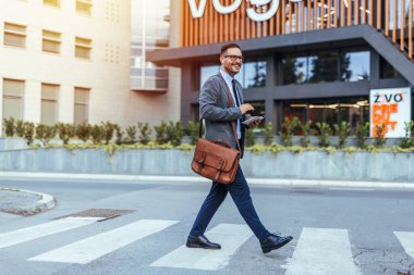 Akıllı bir telefon ve deri bir çantayla karşıdan karşıya geçen kendinden emin bir işadamı. Profesyonel giysiler içinde, şehir ortamında bir başarı ve amaç duygusu yayıyor..