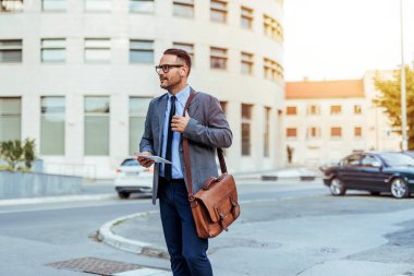 Takım elbiseli kendine güvenen bir işadamı elinde evrak çantası ve belgelerle şehir merkezinde yürüyor. Odaklanmış ve profesyonel görünüyor..