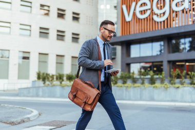 Profesyonel iş adamı, tablet bir cihaz kullanırken bir ofis binasının dışında güvenle yürüyor. Takım elbise giymiş, evrak çantası taşıyor, modern iş hayatı tarzını gözler önüne seriyor..