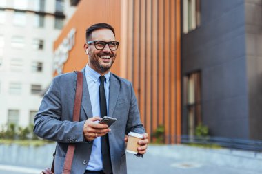 Takım elbiseli kendine güvenen işadamı, elinde kahve ve akıllı telefon tutarken gülümsüyor. Modern iş hayatı ve başarıyı resmediyor..