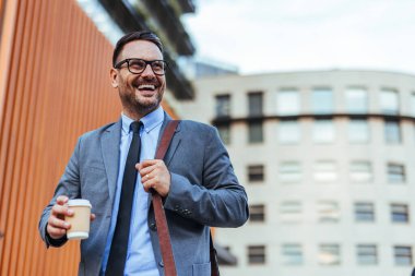 Takım elbiseli gülümseyen iş adamı elinde kahve poşeti, modern bir ofis binasının önünde yürüyor. Kentsel bir ortamda güven, başarı ve profesyonel yaşam tarzını tasvir eden mükemmel bir görüntü..