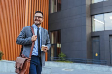 Gülümseyen işadamı elinde kahve fincanı ile takım elbise giyer, modern bir ofis binasının önünde güvenle yürür..