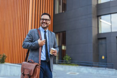 Takım elbiseli, elinde kahve ve deri bir çanta olan güler yüzlü bir işadamı şehir ortamında güvenle yürüyor. Başarı, profesyonel yaşam tarzı ve modern çalışma ortamını temsil eder.