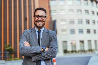 Takım elbise ve gözlük takan kendine güvenen bir işadamı, bir şehir ofis binasının önünde dururken gülümseyerek başarı ve profesyonelliği ifade ediyor..