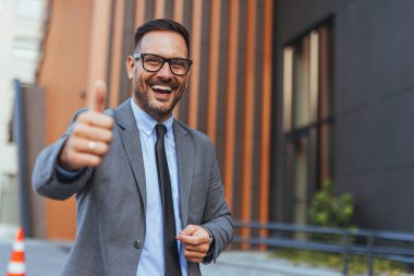Takım elbiseli neşeli bir işadamı, modern bir ofis binasının önünde, profesyonel bir ortamda kendine güven ve pozitiflik sergiliyor..