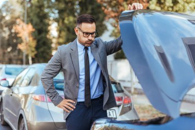 Takım elbiseli endişeli bir işadamı şehir merkezinde bir araba motorunu inceliyor, muhtemelen bir arıza veya mekanik bir sorunla uğraşıyor. Kentsel ortam sorunun çözümünde aciliyet öneriyor.