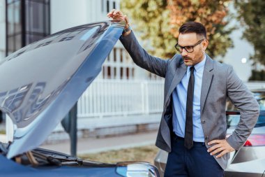 Takım elbiseli endişeli bir işadamı arabasının motorunu inceliyor. Potansiyel sorunları kontrol ediyor, aracında bir sorun var mı diye. Ortam açık bir otoparkta..