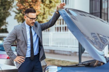 İyi giyimli bir işadamı arabasının motorunu inceler ve endişelerini dile getirir. Ortam, potansiyel bir otomobil arızası veya bakım sorununu gösteren kentsel bir ortam..