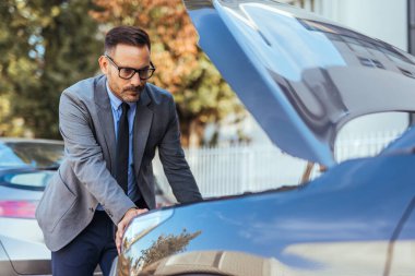 Takım elbiseli bir işadamı kaputu açık bir şekilde arabasının yanında duruyor, açık ve güneşli bir günde motoru inceliyor..