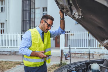 Güvenlik yeleği giyen bir adam, akıllı telefon kullanarak bir araba motorunu endişeyle inceliyor. Sahne, yol kenarı sorununa işaret eden kentsel bir zemin üzerine kurulmuş..