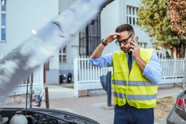 Yansıtıcı yelek giyen endişeli bir adam şehir caddesinde kaputu açık bozuk arabasının yanında bir telefon görüşmesi yapar..
