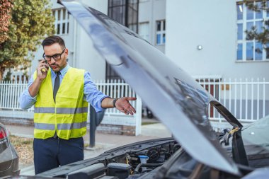 Emniyet yeleği giyen bir adam araba motorunu incelerken bir telefon görüşmesi yapıyor. Olay yerinde bir araç arızası veya bakım durumu olduğu öne sürülüyor, endişe ve tedbir vurgulanıyor.
