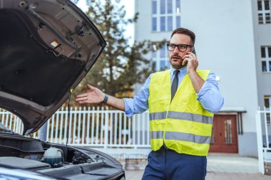 Güvenlik yeleği giyen bir adam bozuk arabasının yanına yardım çağırmak için cep telefonu kullanıyor. Arabanın kaputu açık ve endişeli görünüyor..