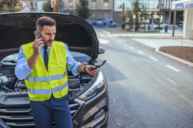 Emniyet yeleği giymiş endişeli bir adam, şehir merkezinde bozuk arabasının yanında yardım çağırmak için telefonunu kullanıyor..