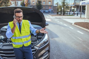 Yansıtıcı yelek giyen bir adam bozuk bir arabanın yanında duruyor ve bir şehir caddesinde yol yardımı için telefon ediyor. Endişeli ve sinirli görünüyor..