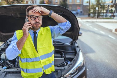 Emniyet yeleği giymiş hayal kırıklığına uğramış bir adam açık kapüşonlu arabasının yanında bir telefon görüşmesi yapıyor. Endişeli görünüyor ve yol kenarında duruyor, yol yardımı istiyor..