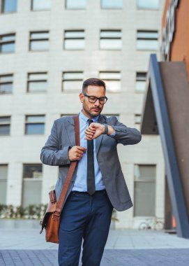 Takım elbiseli bir iş adamı şehir ortamında yürürken saatini kontrol eder. Modern şehir yaşamında dakikliği ve organizasyonu simgeleyen deri bir çanta taşıyor..