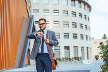 İyi giyimli bir işadamı modern bir ofis binasının önünde durmuş saatini kontrol ediyor. Görüntü, kurumsal bir ortamda profesyonellik, dakiklik ve kentsel yaşam tarzının temalarını aktarır..