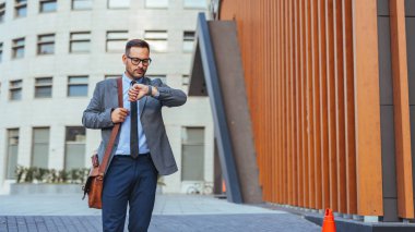 Takım elbiseli profesyonel bir adam modern bir binanın önünden geçerken şehir ortamında dakiklik ve iş hayatı tarzını kontrol eder..