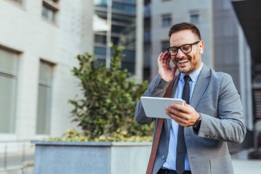 Şehir ortamında tablet ve kulaklıkları olan neşeli bir iş adamı. Bağlılık ve modern iş kültürü sergiliyor. Takım elbise giymiş, meşgul ve açık havada üretken görünüyor..