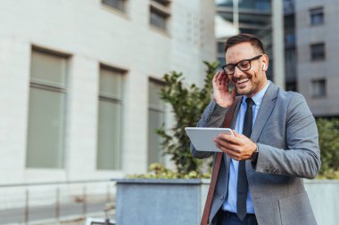 Neşeli bir iş adamı, bir şehirde dışarıda dururken tablet üzerinde video aramasından zevk alır. Gülümsüyor ve gözlük takıyor, profesyonellik ve modern bağlılık yayıyor..