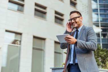 Modern bir ofis binasının önünde kulaklık takarken tablet kullanan takım elbiseli gülümseyen işadamı, iş dünyasında teknoloji ve iletişimi temsil ediyor..