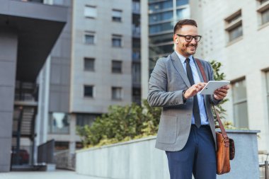Takım elbiseli kendine güvenen bir işadamı çağdaş bir ofis binasının yakınında açık havada tablet kullanıyor. Odaklanmış ve profesyonel görünüyor, modern kentsel iş hayatı hissi yayıyor..