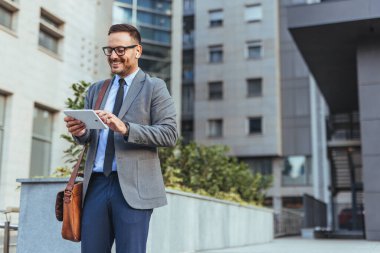 Gülümseyen işadamı, kulaklık takarken tablet kullanarak dışarıda duruyor. Profesyonel olarak giyinmiş, özgüvenini ve modern teknoloji kullanımını ifade ediyor. Kentsel arka plan bir şehir iş ortamı öneriyor.