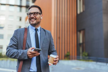 Takım elbiseli kendine güvenen bir işadamı, elinde akıllı telefon ve kahveyle dışarıda gülümsüyor. Şehir ortamında modern iş hayatı, başarı ve bağlantıyı temsil eder. Kariyer, iletişim ve teknoloji temaları için ideal.