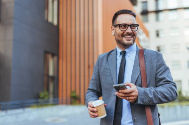 Takım elbiseli, akıllı telefonunu kullanan ve elinde kahve fincanı tutan neşeli bir işadamı gülümseyen ve pozitif bir tavır sergileyen modern bir şehir bölgesinde güvenle yürüyor..